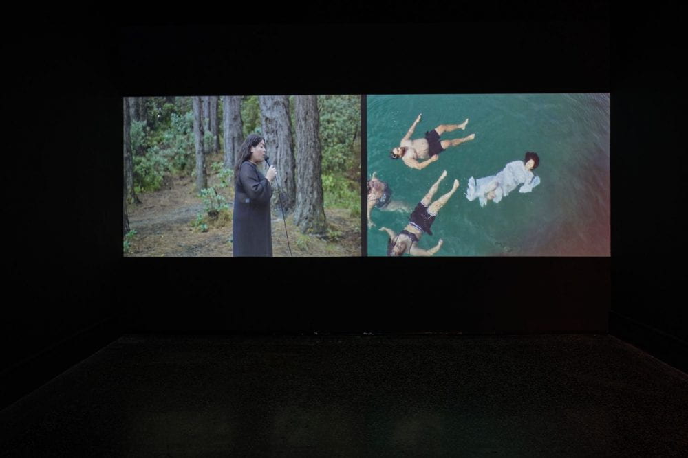 A two-channel film projected in a dark room. In the left film a woman in a black dress sings in a forest. In the right-hand film, three people float on their backs in the ocean.