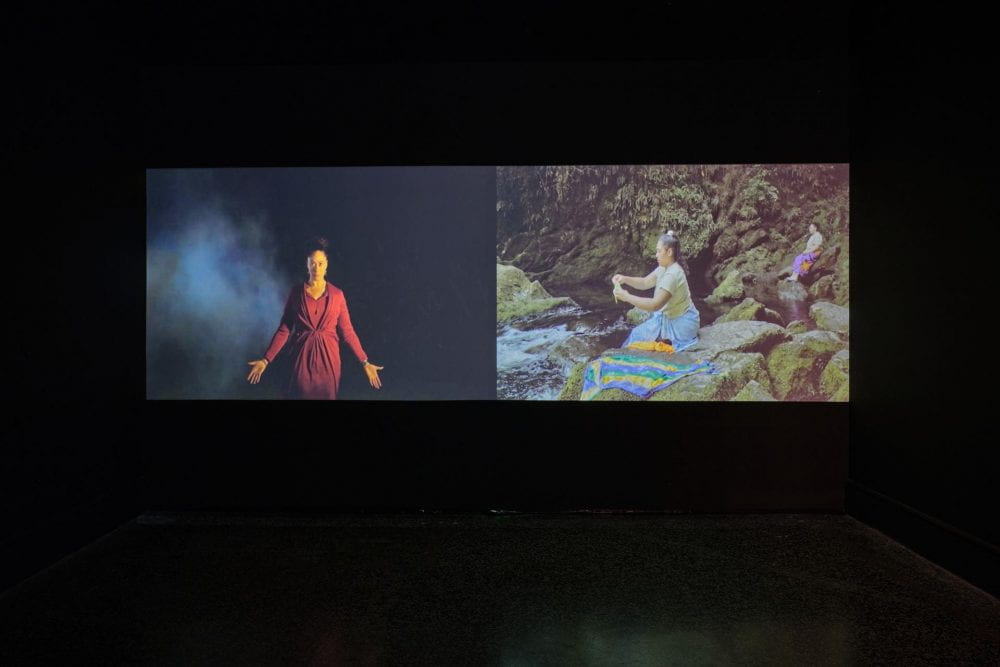 A two-channel film projected in a dark room. On the left, a woman in red stares at the camera with her arms outstretched. In the right-hand film, a woman sits on a rock in a riverbed.