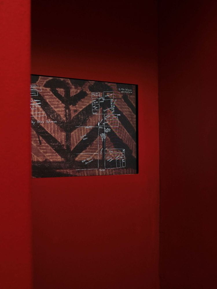 A booth space painted red, with a screen embedded in the back wall. On the screen, the film shows a hand-drawn map of a house layout against a brown and black tapa cloth.