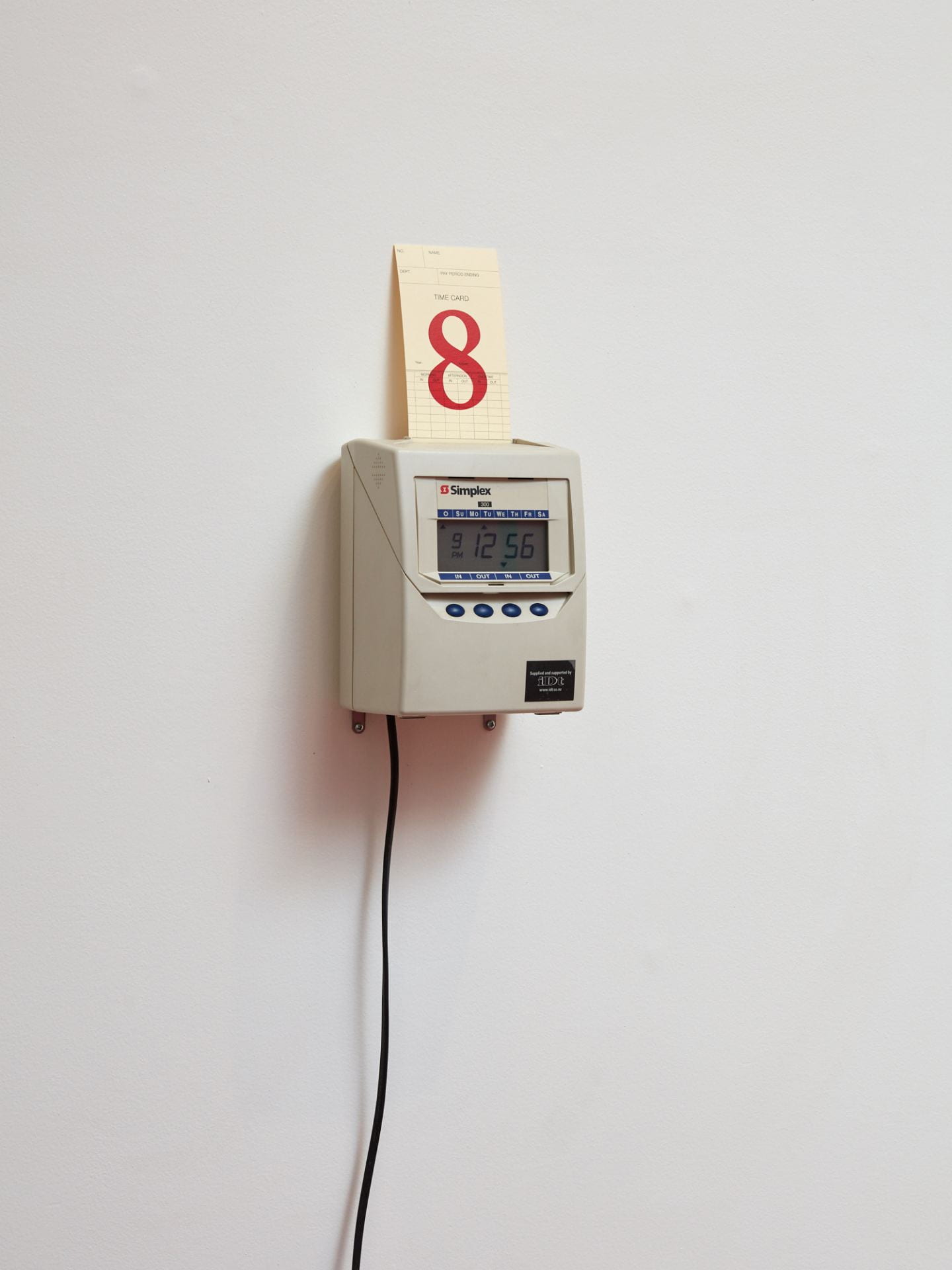 An old-fashioned employee timeclock mounted on a gallery wall. A timecard rests in the slot on top, with a red '8' stamped on it.
