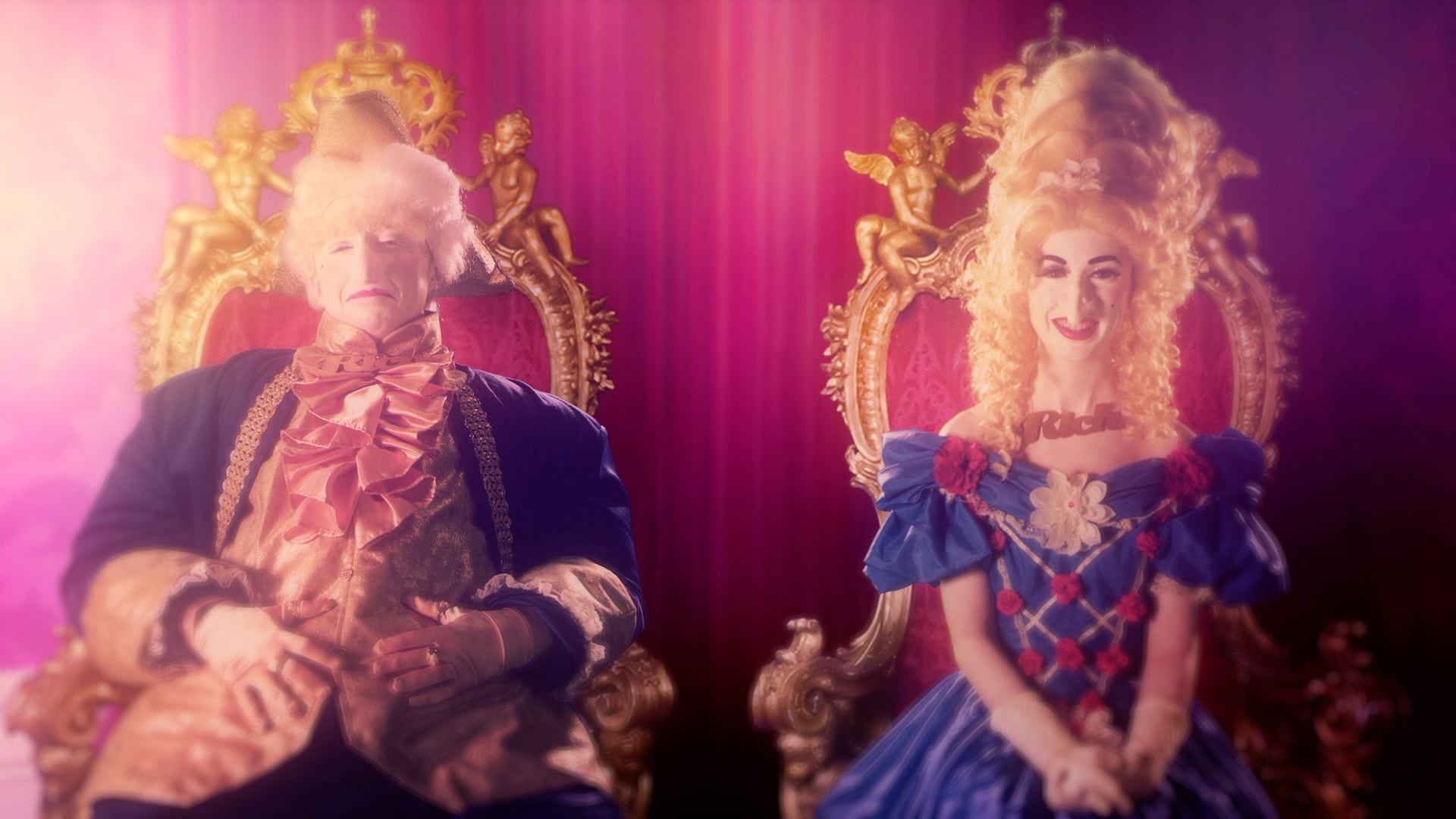 A king and queen sit side by side on matching thrones. They are dressed in exaggerated, garish Georgian outfits with powdered wigs and prosthetic facial features.