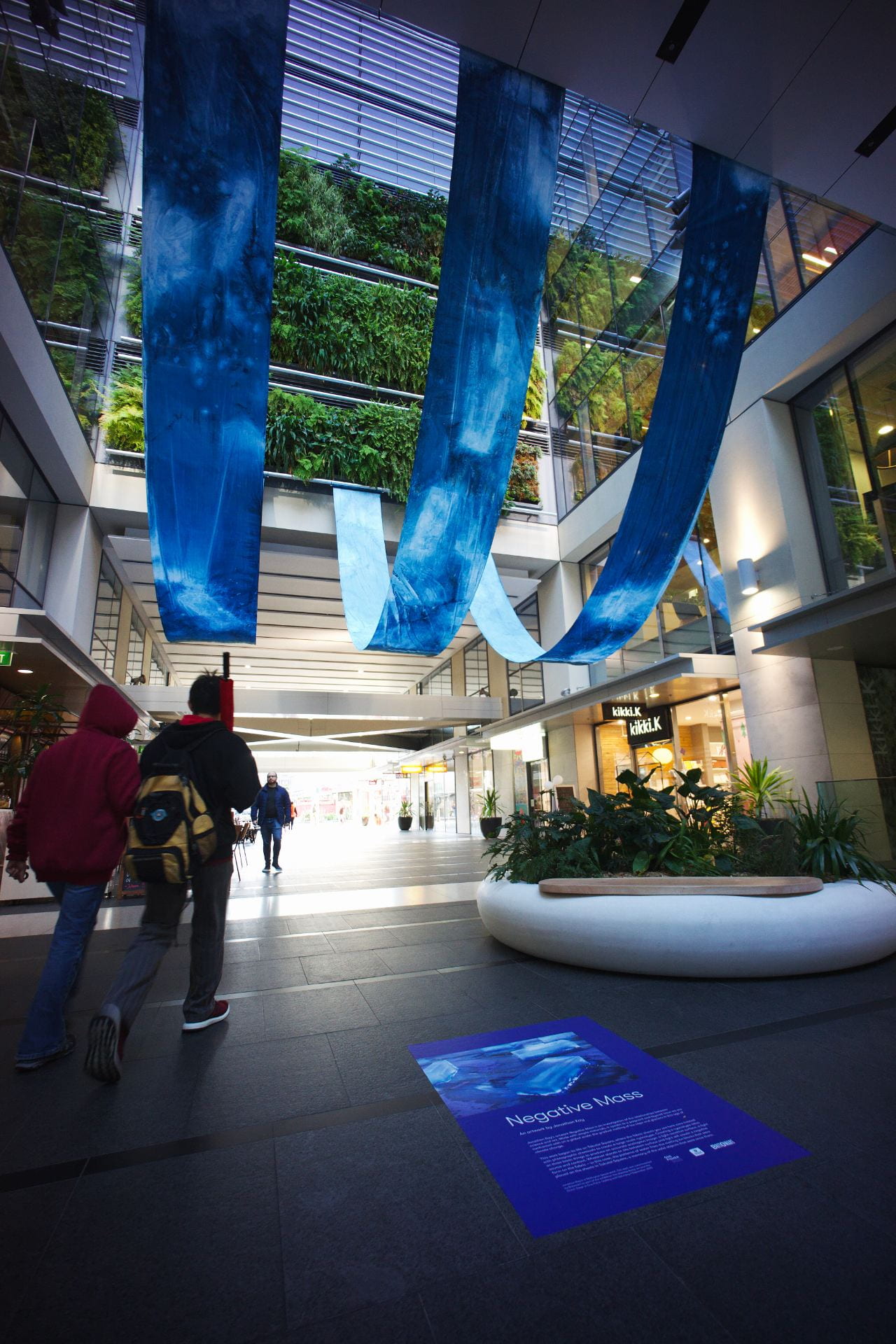 Three long, narrow strips of cloth hang in U-shapes in the courtyard of an inner-city building. A living plant wall can be seen in the background.