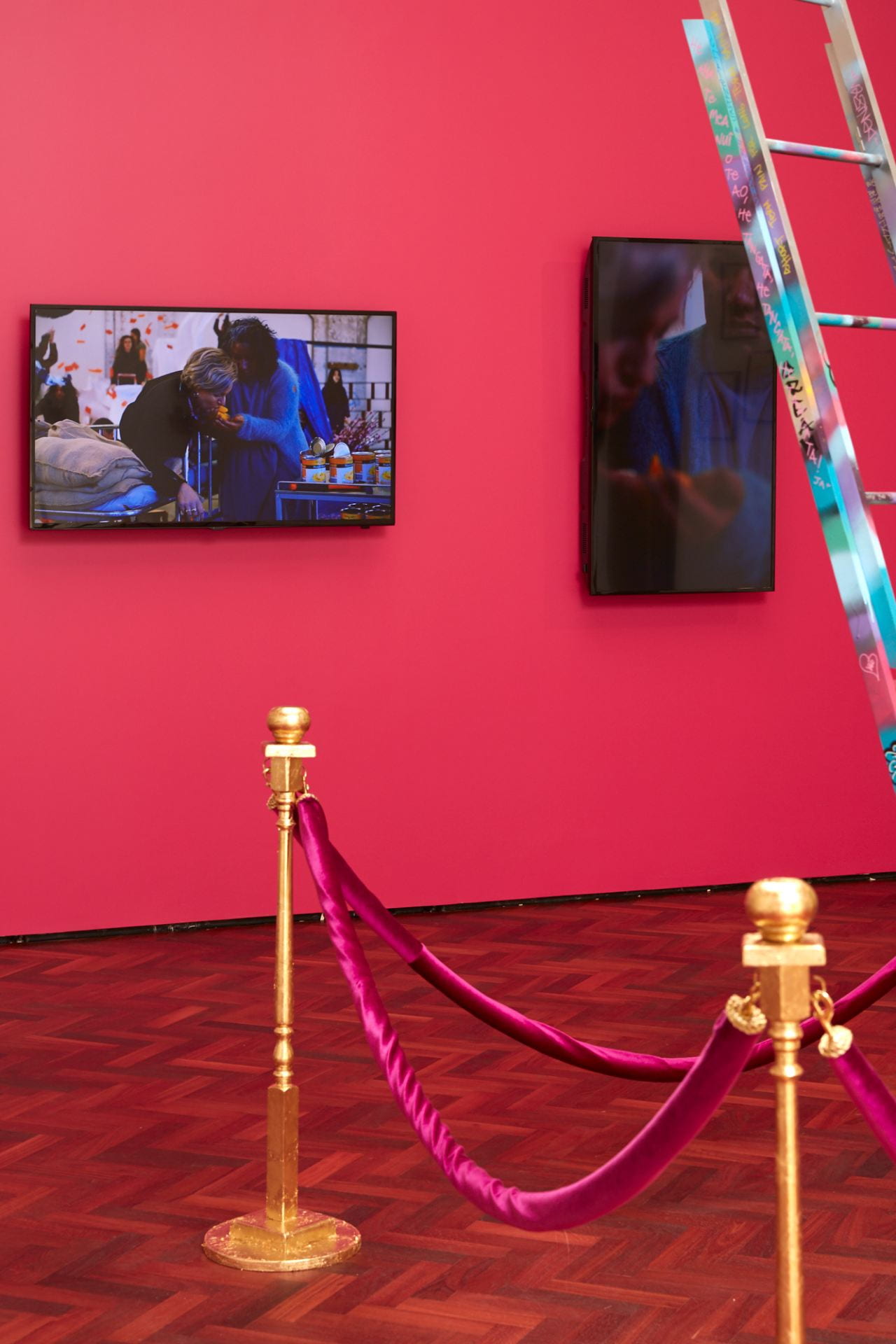 A hot pink wall with two TVs mounted on it, showing a silent montage of people in the throes of chaos, throwing packaged food, feeding one another and setting paper on fire.
