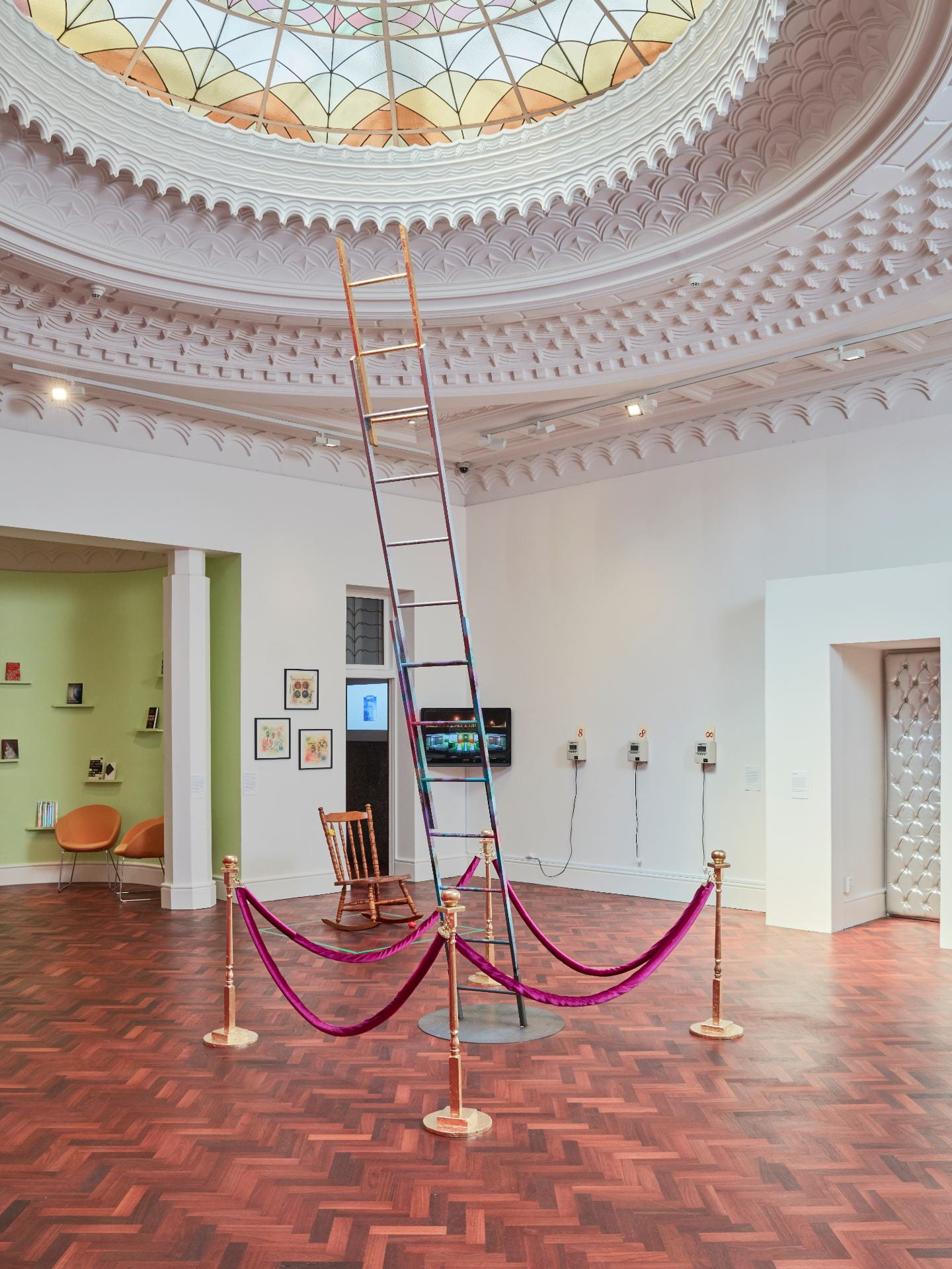 A gallery space with an elaborate dome ceiling and wooden floor. In the centre of the room a ladder decorated with graffiti and gilded at the top stretches towards the ceiling, surrounded by gold bollards with velvet pink ropes.
