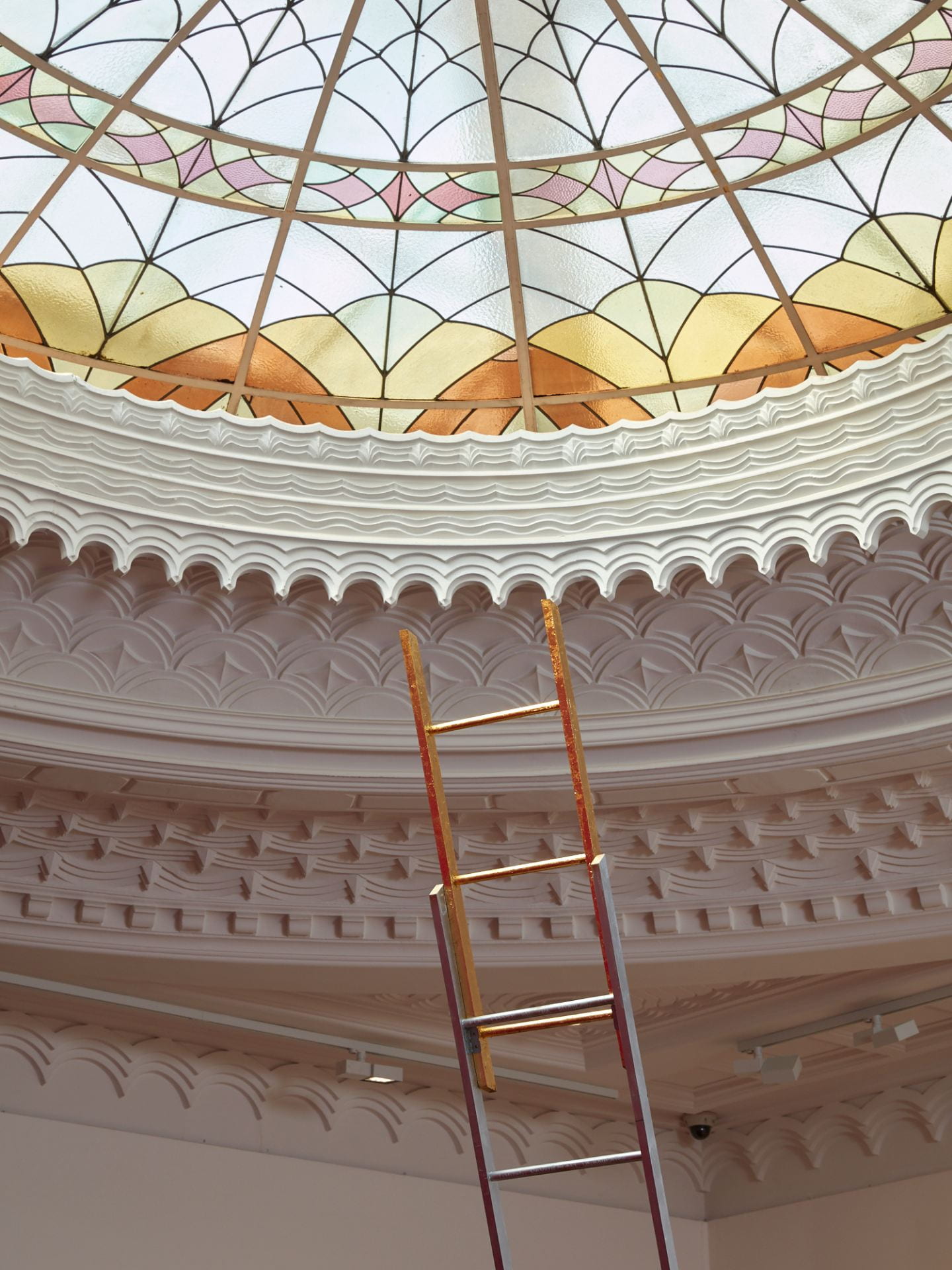 The gilded top of a ladder rises up towards the ceiling. The ceiling is an elaborate stained glass dome, with patterned plaster moulding around the outside.