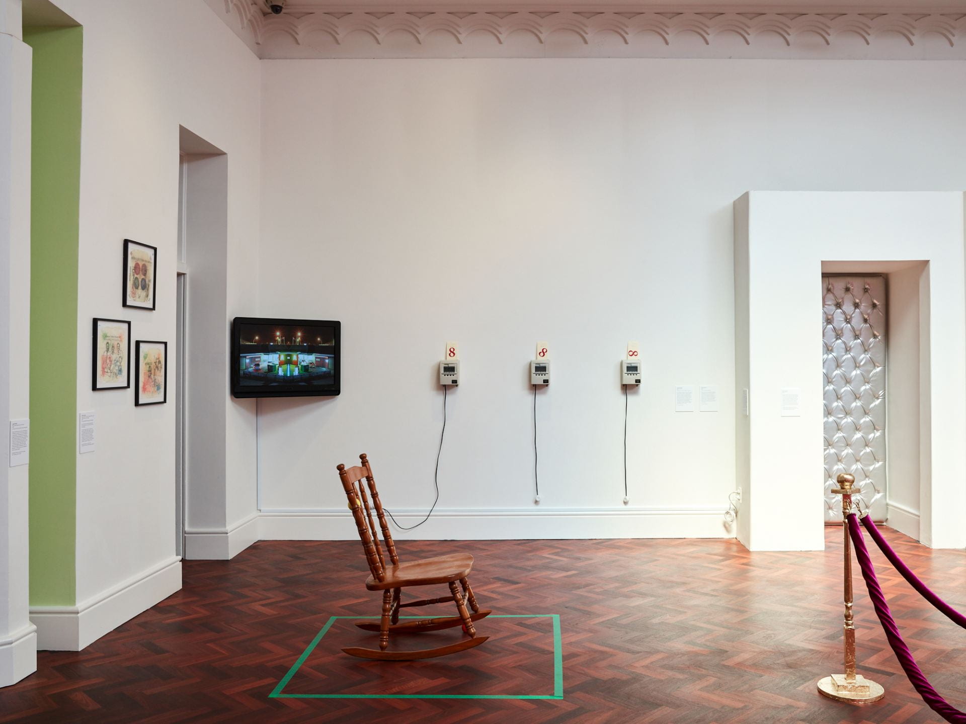 A gallery space with white walls and wooden floor. A rocking chair sits on the floor, a stress ball wedged under one rocker. A TV, three timestamp clocks and a booth space are all visible in the background.