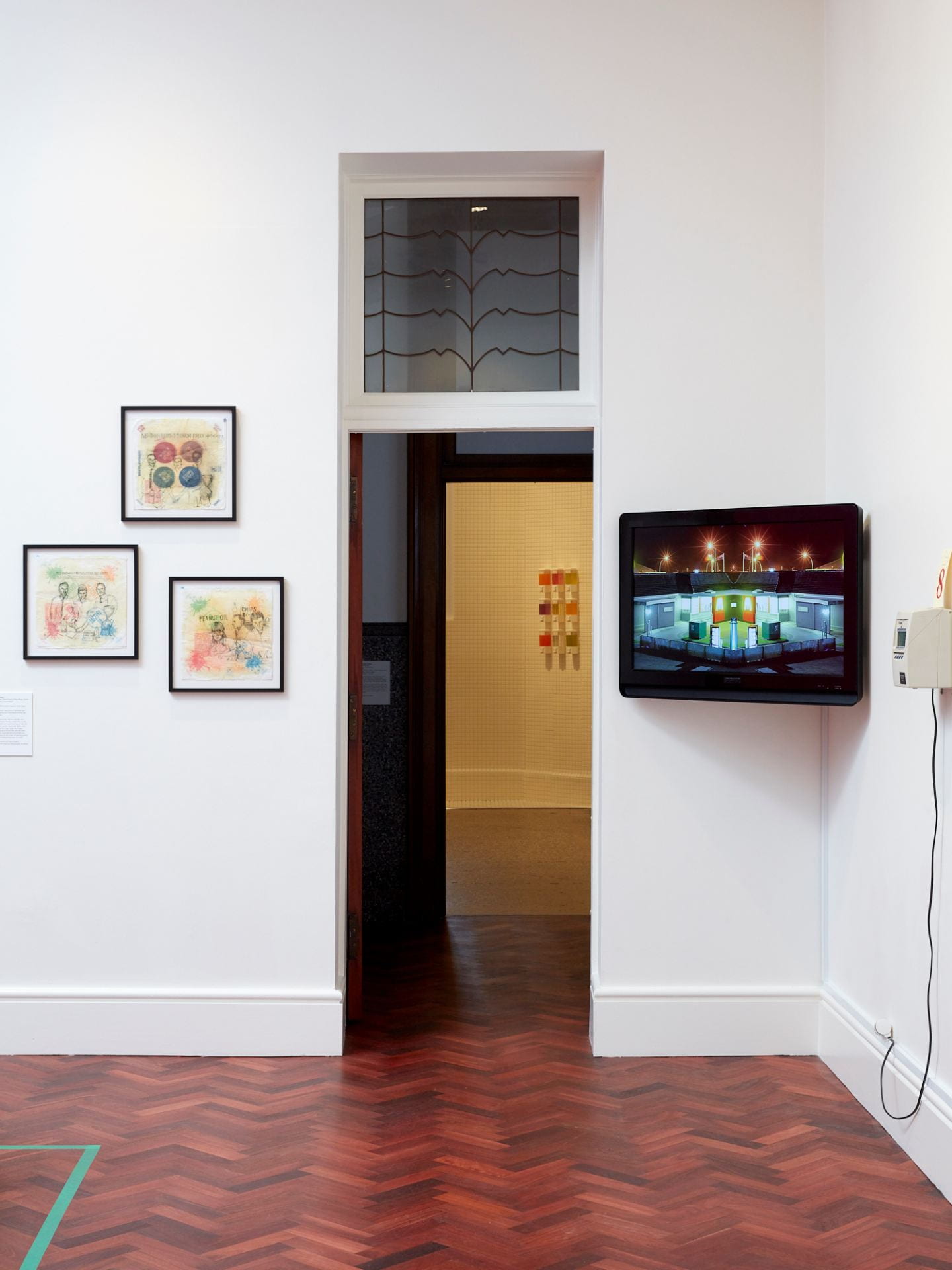 A door into another gallery. On the left of the door is three framed artworks of McDonald's wrappers with ink drawings traced onto each. On the right is a flatscreen TV, mounted against the corner of the wall, showing a car endlessly looping through a McDonald's drive thru.