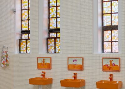 A gallery space covered in white tiles, with coloured vinyl applied over three tall windows and three orange sinks with elephant taps mounted to the wall. Above each sink is a painting of the same sinks.