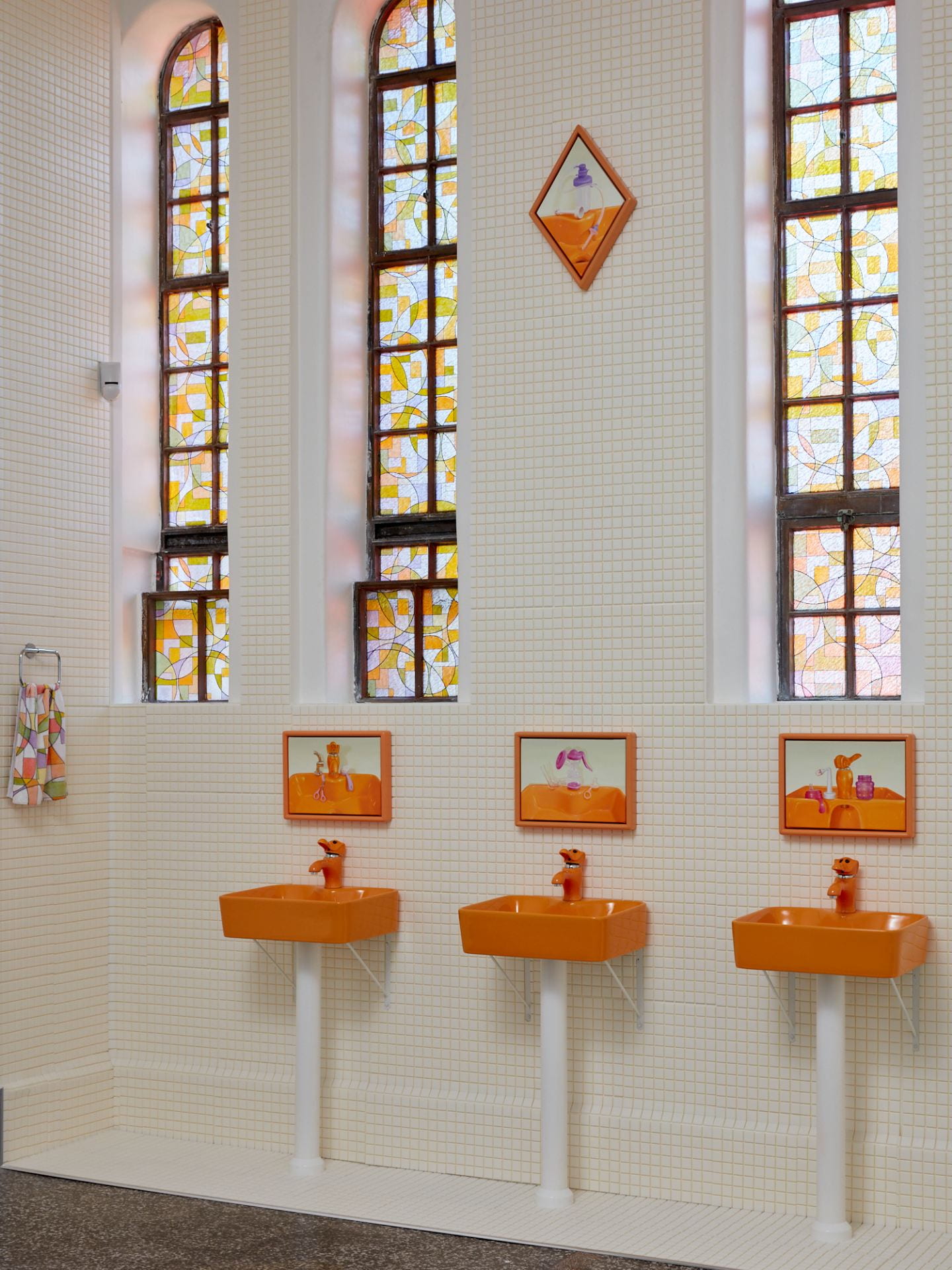 A gallery space covered in white tiles, with coloured vinyl applied over three tall windows and three orange sinks with elephant taps mounted to the wall. Above each sink is a painting of the same sinks.