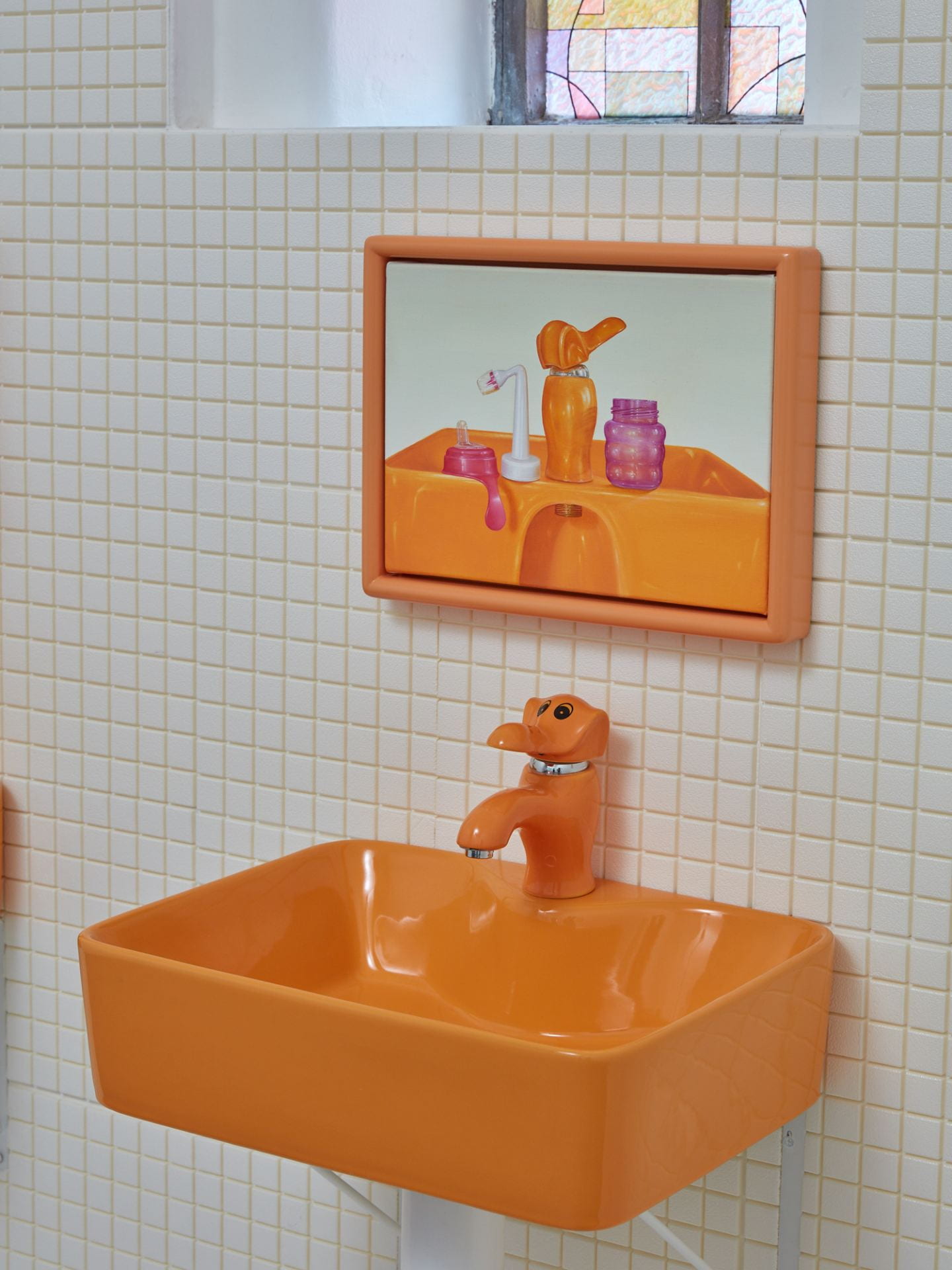 An orange sink with an elephant shaped tap installed on a white tiled wall. Above the sink is a small painting of the sink itself.
