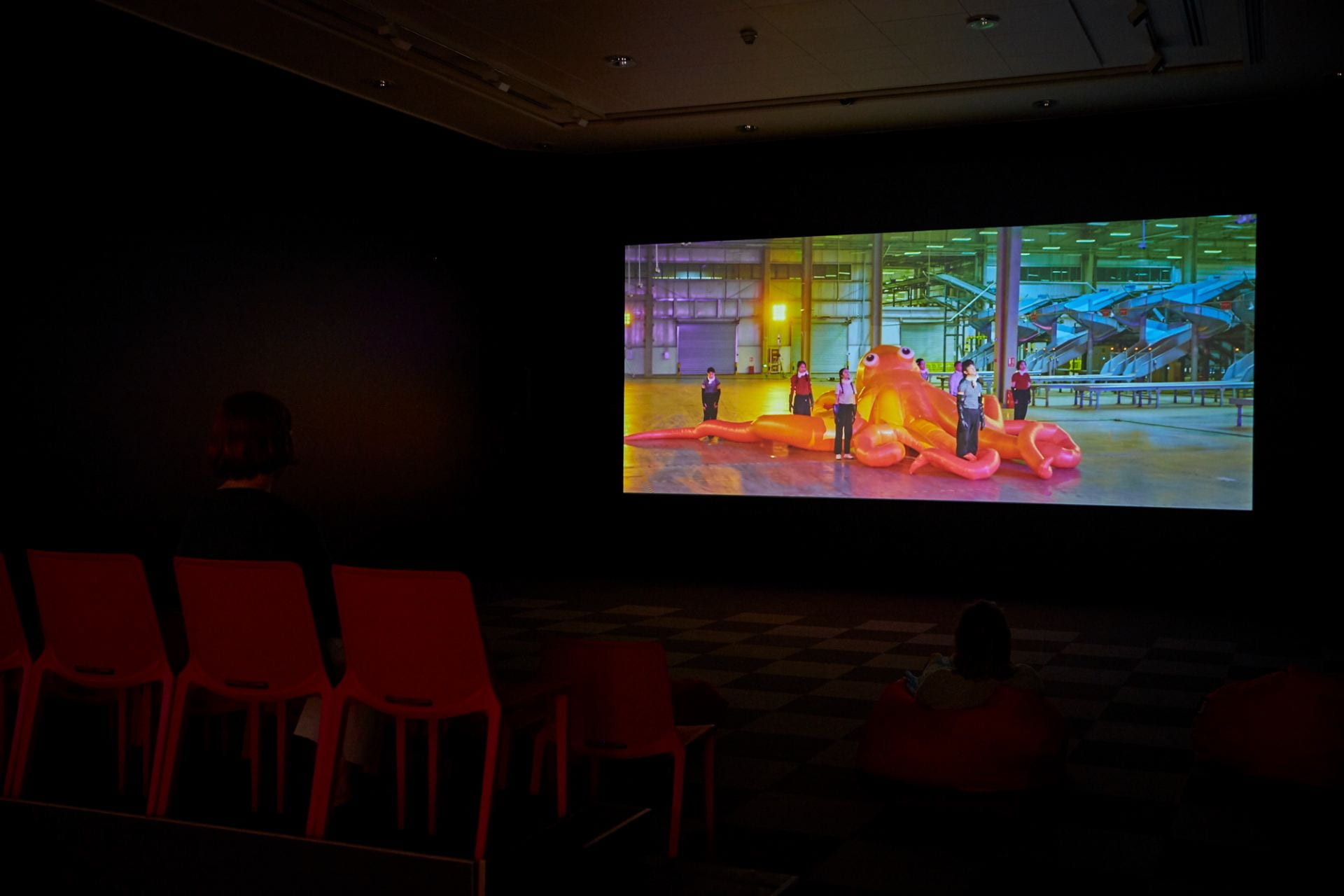 A film projected in a dark room. In the film, a huge inflated bright orange octopus is surrounded by dancers standing among its tentacles. The scene takes place in a dimly lit warehouse.
