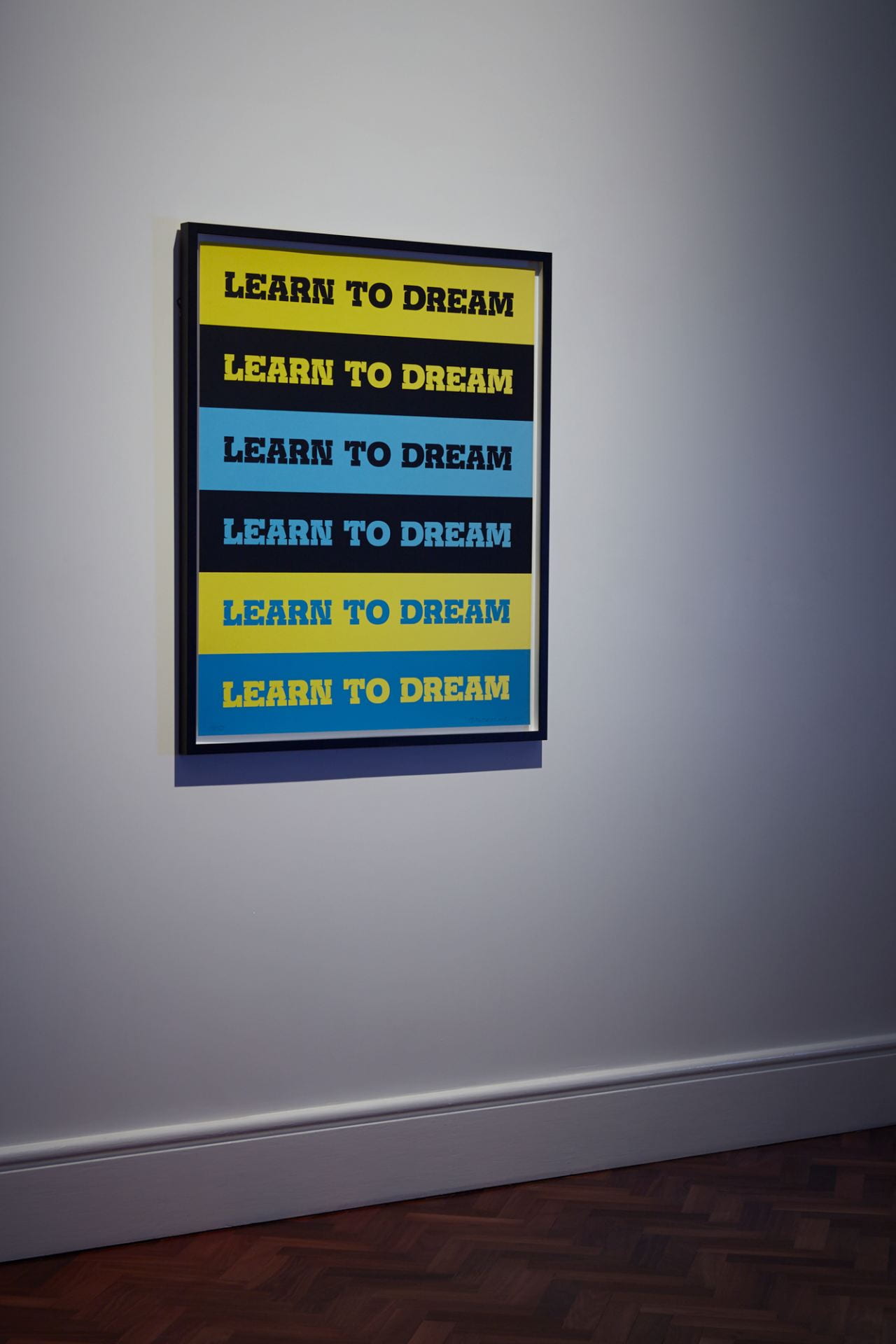 A framed artwork made up of yellow, black and blue horizontal stripes. Each stripe has the phrase "Learn to dream" written across it.