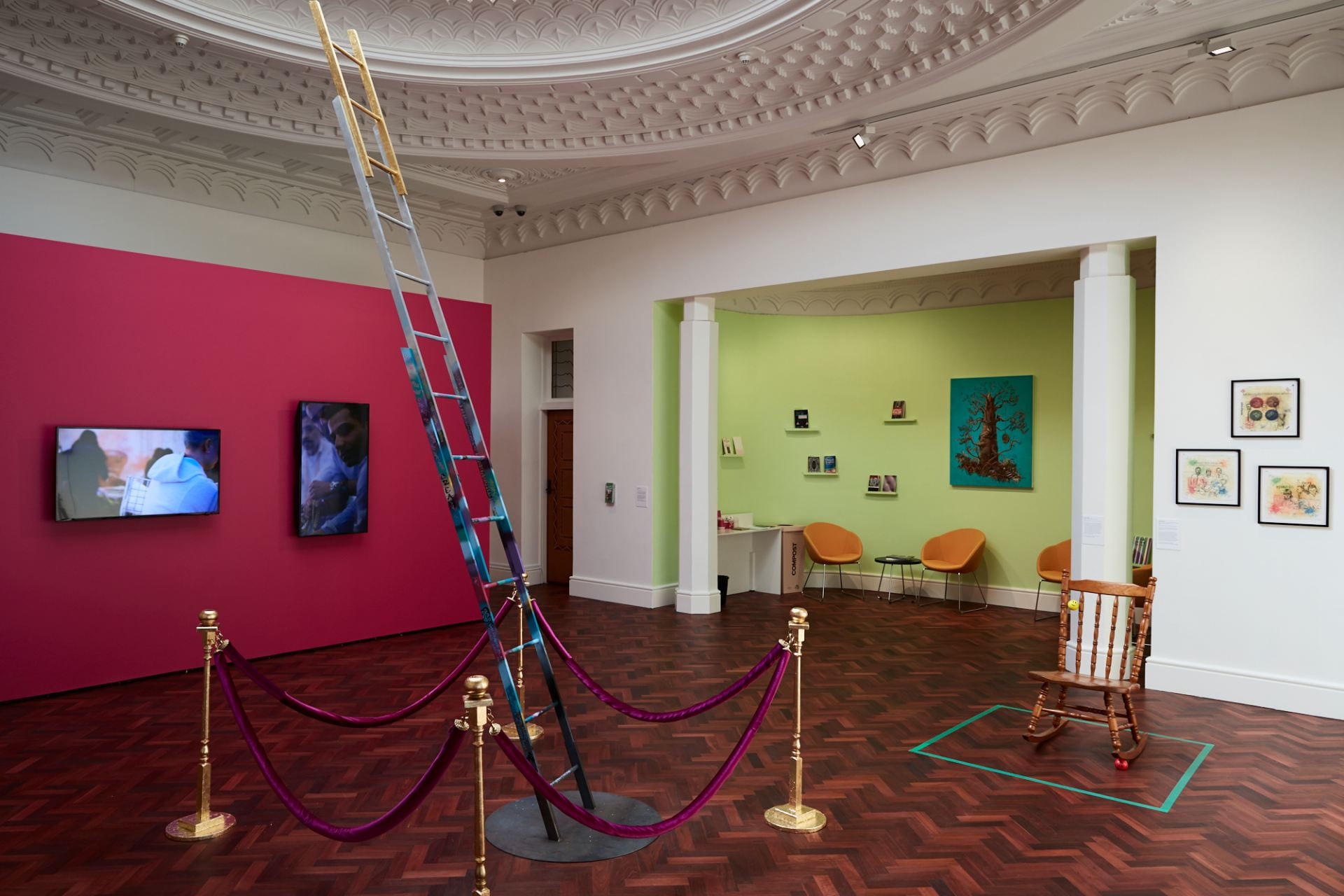 A gallery space with an elaborate dome ceiling and wooden floor. A hot pink wall in the background has two televisions hung on it. In the foreground, a ladder decorated with graffiti and gilded at the top stretches towards the ceiling, surrounded by gold bollards with velvet pink ropes.