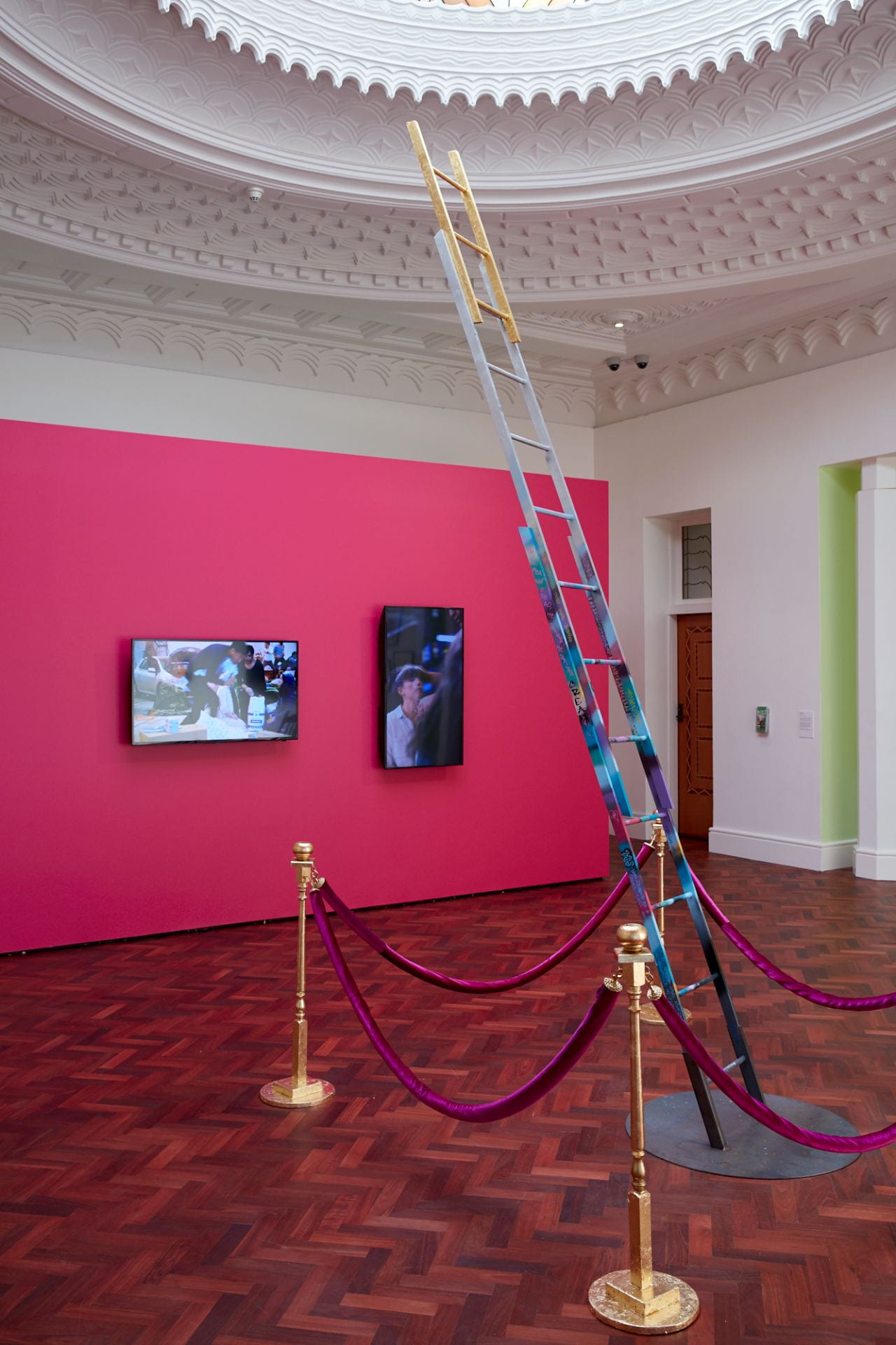 A gallery space with an elaborate dome ceiling and wooden floor. A hot pink wall in the background has two televisions hung on it. In the foreground, a ladder decorated with graffiti and gilded at the top stretches towards the ceiling, surrounded by gold bollards with velvet pink ropes.