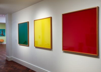 Three framed squares of green, yellow and red, hung along a white corridor wall.