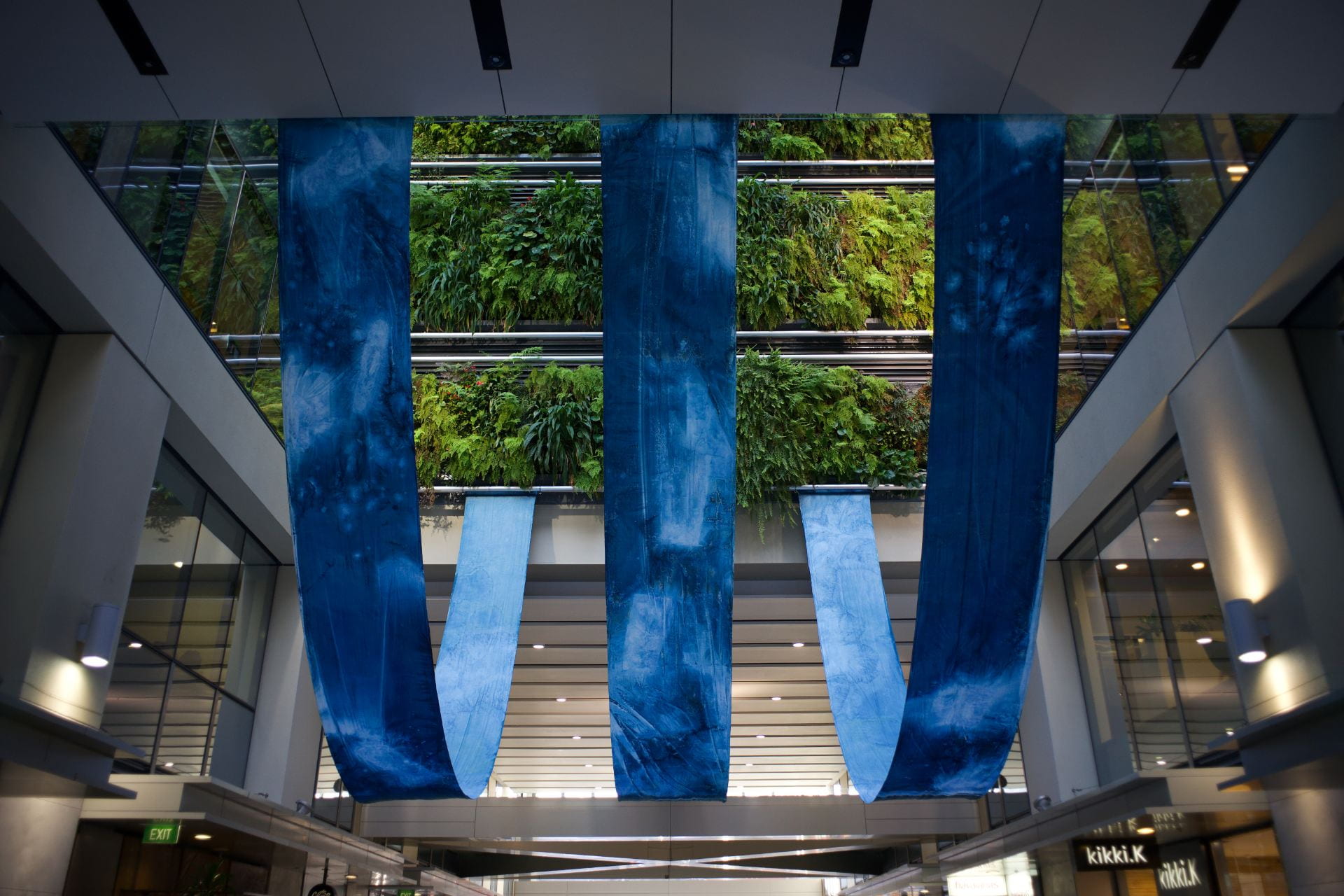 Three long, narrow strips of cloth hang in U-shapes in the courtyard of an inner-city building. A living plant wall can be seen in the background.