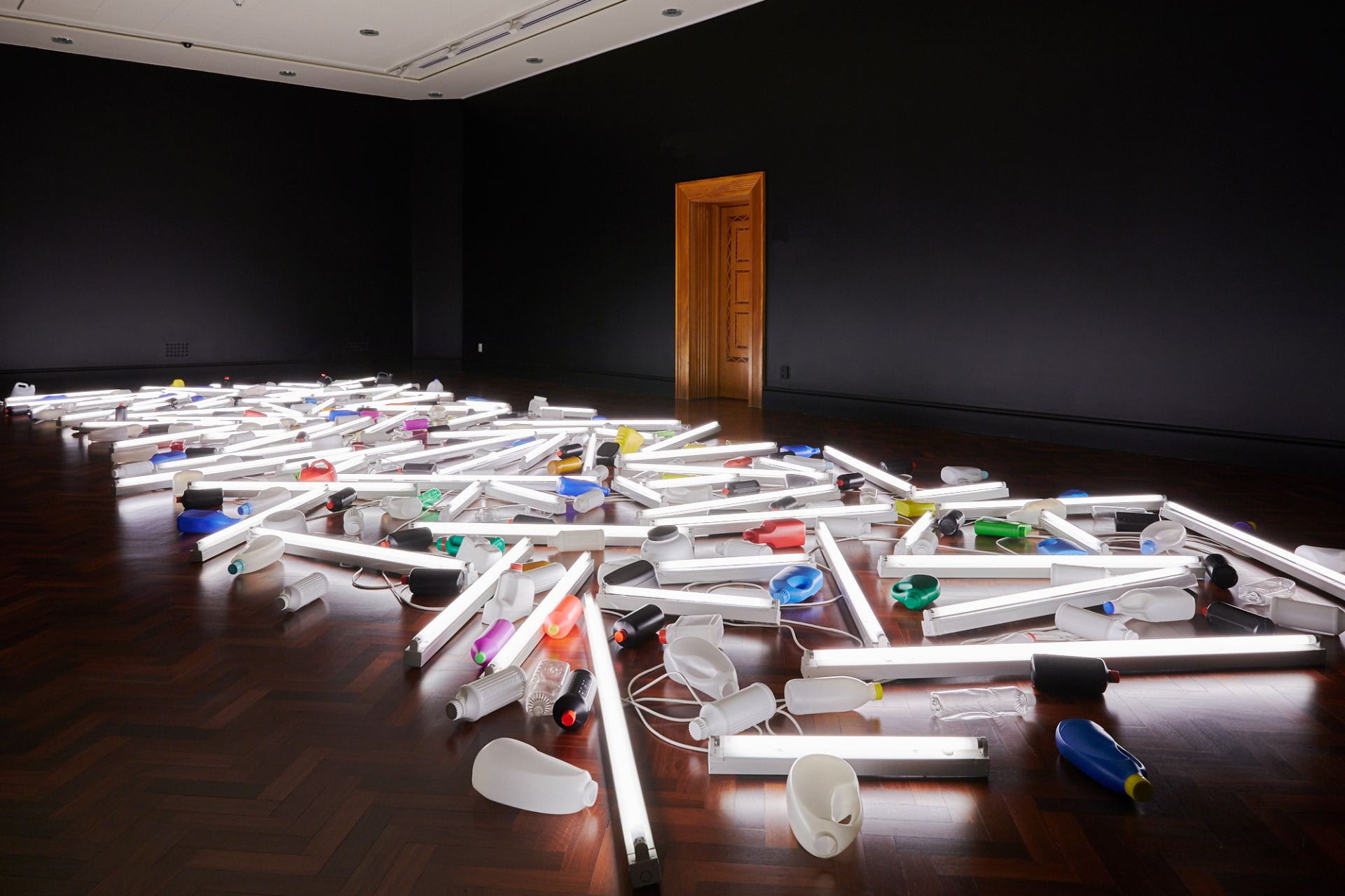 A long, wide assortment of plastic bottles and tubular fluorescent lights are arranged diagonally on a wooden floor, illuminating the room.