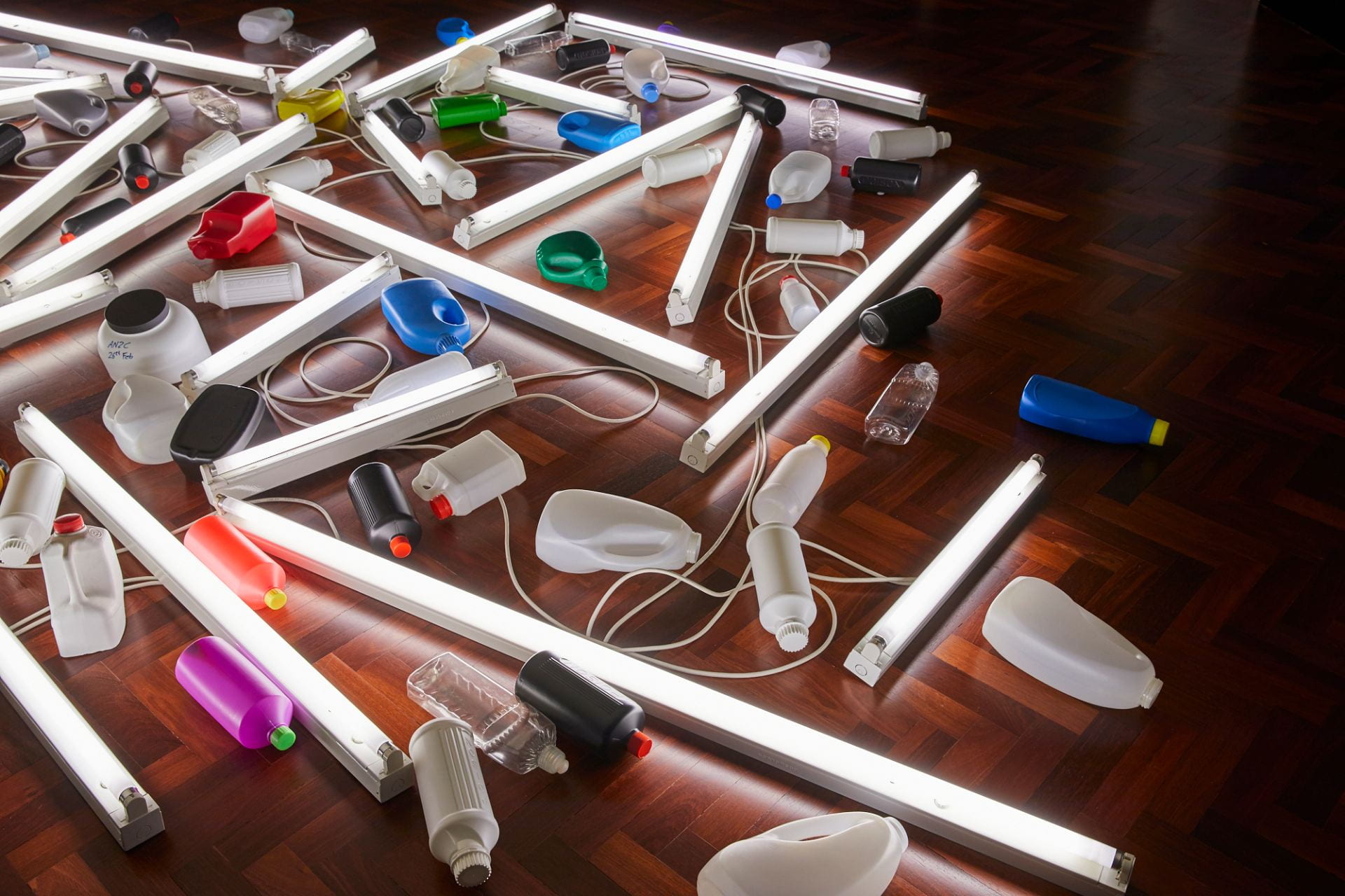Close-up of an assortment of plastic bottles and tubular fluorescent lights arranged diagonally on a wooden floor.