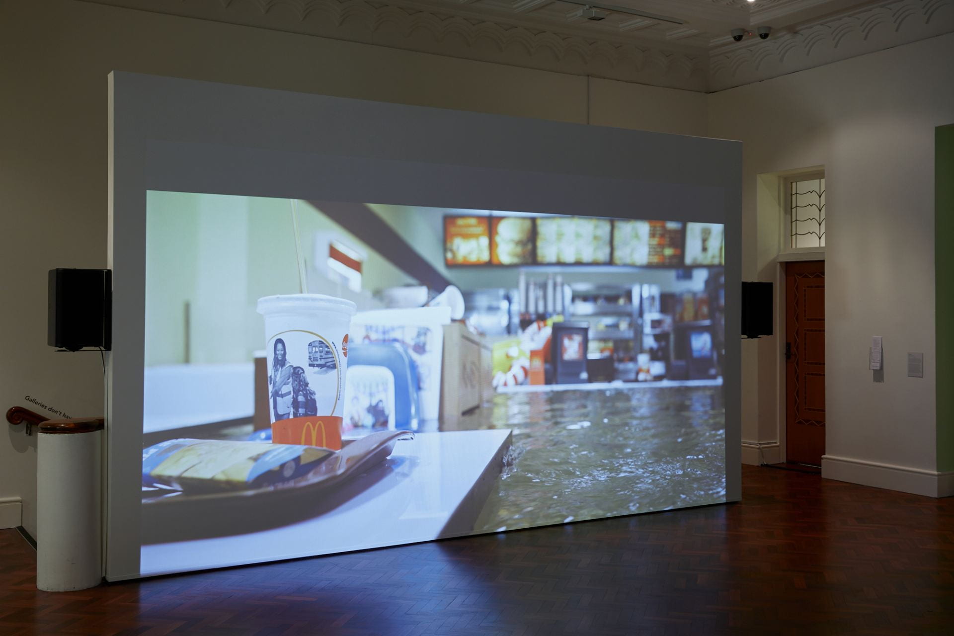 A projection screen shows a film. In the film, a McDonald's restaurant is being slowly filled with water. The water is at counter height.