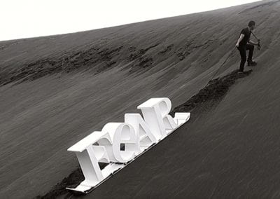 A man drags a white sled up a tall black sand dune. The sled has giant 3D white letters on it spelling 'FEAR'
