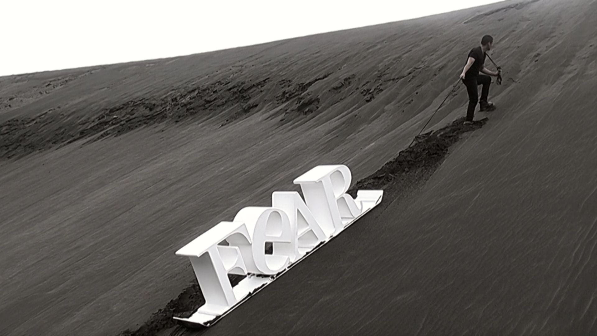A man drags a white sled up a tall black sand dune. The sled has giant 3D white letters on it spelling 'FEAR'