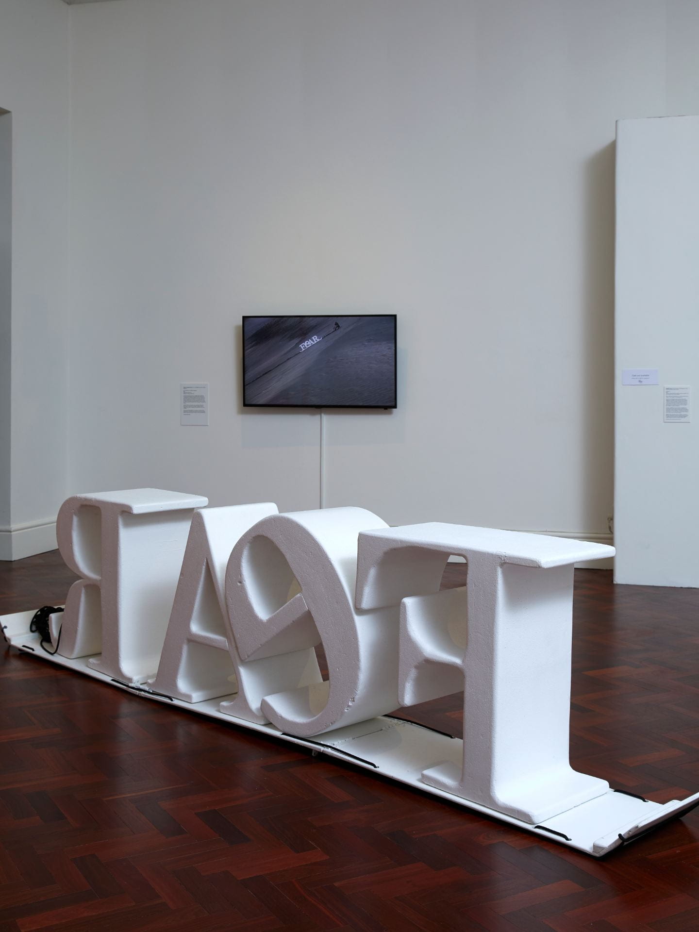 A 3D sculpture of giant white letters spelling out 'FEAR', with a TV playing an accompanying film visible on the wall in the background.