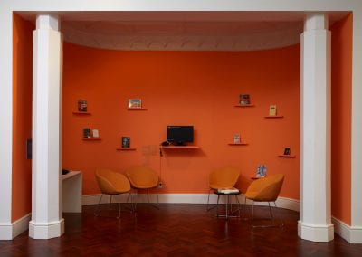An alcove space painted orange, with chairs and books for reading. A computer monitor sits in the middle playing recordings.