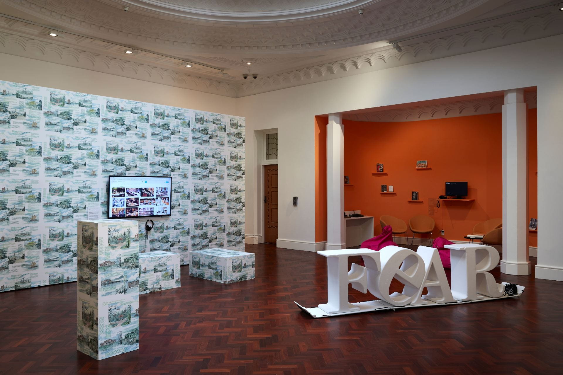 A gallery room. On the left is a wall covered in wallpaper depicting landmarks around Auckland. On the right is an alcove painted orange, and giant white 3D letters spelling out 'FEAR'.