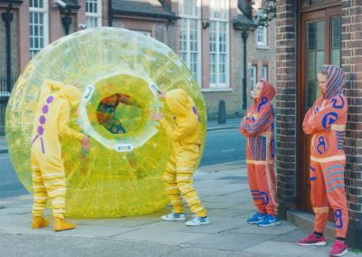 A person in a yellow inflatable zorb ball attempts to gain access to a bar, guarded by two people dressed in orange and blue onesies. The overall scene parodies the way viruses stealthily enter and infect a person's immune system.