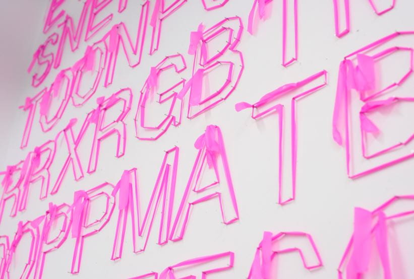 A narrow booth space filled with letters. Each letter is constructed from pink trail tape stretched around nails. Overall it looks like a word search.