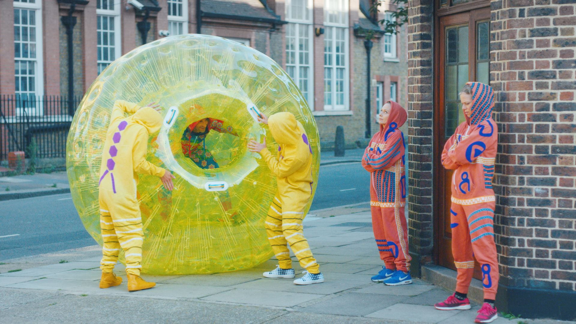 A person in a yellow inflatable zorb ball attempts to gain access to a bar, guarded by two people dressed in orange and blue onesies. The overall scene parodies the way viruses stealthily enter and infect a person's immune system.