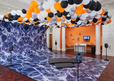 A huge wave structure surges up in the space, with a sea of orange, black, silver and white balloons hanging overhead. Four speakers are positioned around the wave, with acoustic foam cushions in the middle for sitting on.