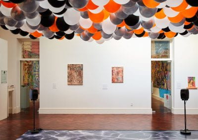 Two abstract pink and grey paintings hang on a white gallery wall. A sea of orange, black, silver and white balloons hang overhead.