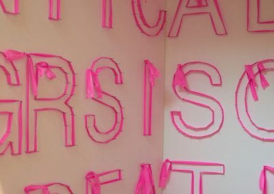 A narrow booth space filled with letters. Each letter is constructed from pink trail tape stretched around nails. Overall it looks like a word search.