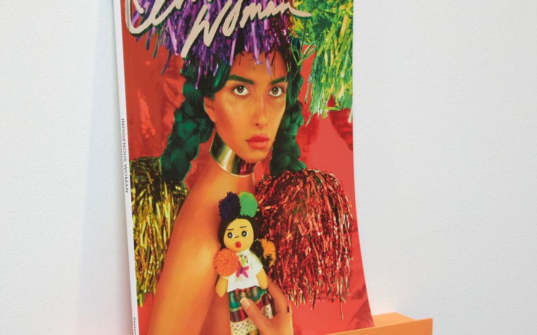 A magazine sitting on an orange bookshelf. The magazine has a woman on the front of it, and its title "Indigenous Woman".
