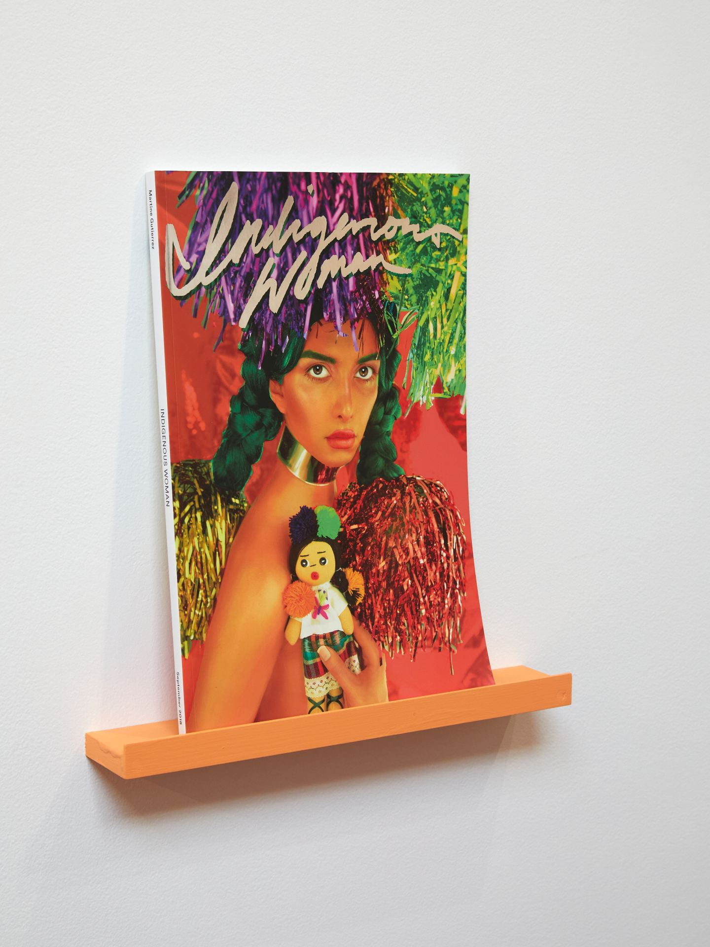 A magazine sitting on an orange bookshelf. The magazine has a woman on the front of it, and its title "Indigenous Woman".