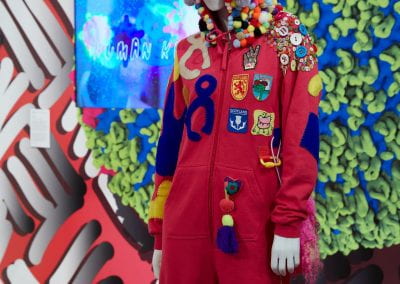 A mannequin dressed in a hot pink onesie, with a hood covered in pom-poms and Scotland-themed badges and abstract blue, grey and yellow shapes sewn to the body.