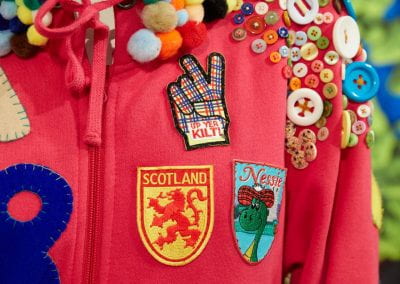 Close-up of a mannequin dressed in a hot pink onesie, with a hood covered in pom-poms and Scotland-themed badges and abstract blue, grey and yellow shapes sewn to the body.