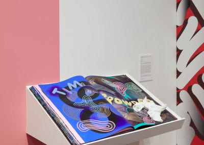 A large book sitting on a slanted shelf. The pages of the book are covered with artworks of abstract shapes and letters on bright colorful backgrounds.