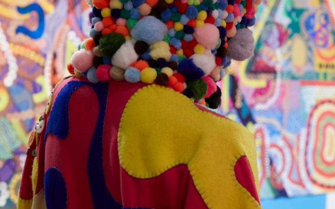 Close-up of the back of a mannequin wearing a hot pink onesie, with a hood covered in pom-poms and yellow and blue patches sewn onto the body.