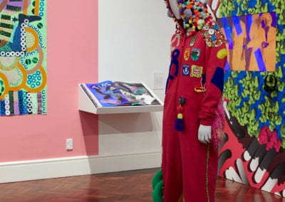 A mannequin dressed in a hot pink onesie, with a hood covered in pom-poms and Scotland-themed badges and abstract blue, grey and yellow shapes sewn to the body. The mannequin stands in a gallery space, with an open artist's book and a TV playing a film in the background.