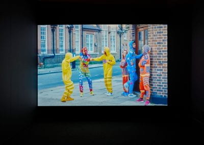 A person in a rainbow coloured onesie holds hands with two people in yellow onesies, outside a bar entrance. The bar entrance is guarded by two people in blue and orange onesies, and they are letting through a person in a blue onesie. This parodies the way viruses enter a human body's immune system by stealth.