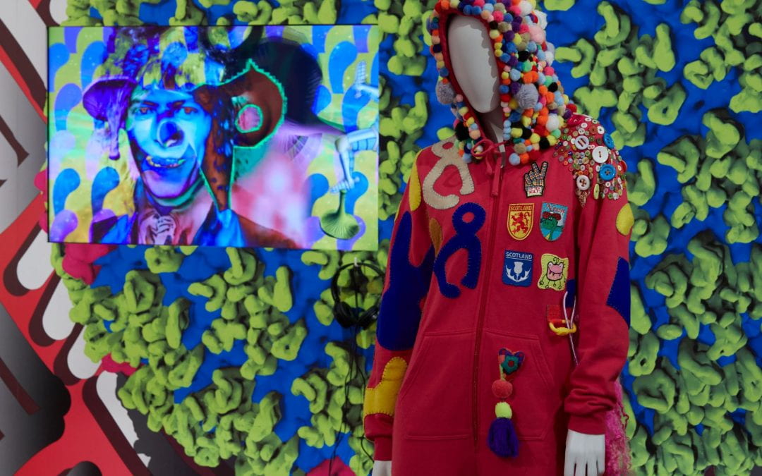On the left is a TV screen playing a psychedelic film, showing a man's face against a colourful background. On the right is a mannequin dressed in a hot pink onesie, with a hood covered in pom-poms and Scotland-themed badges and abstract blue, grey and yellow shapes sewn to the body.