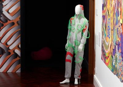 A mannequin stands in a gallery space dressed in a grey onesie with abstract red squiggles and transparent green tubing sewn all over it.