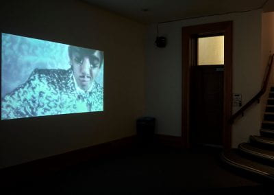 A film projected onto a wall in a dark space. The film shows a person with a moustache and embroidered suit dancing.
