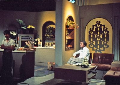 A man sits in a television set styled to look like a 1970s home interior. He sits in the living room, adjacent to an open plan kitchen.