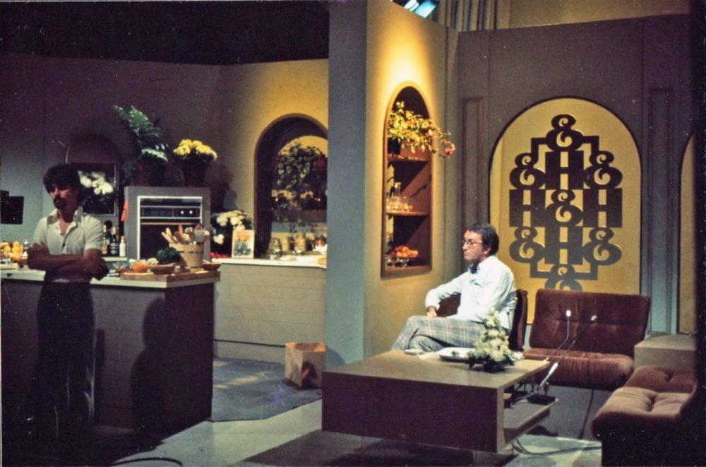 A man sits in a television set styled to look like a 1970s home interior. He sits in the living room, adjacent to an open plan kitchen.