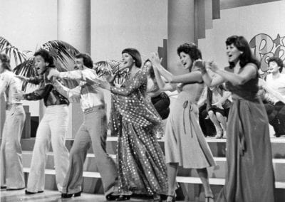 Dancers in flowy dresses and pants groove on a television set, styled to look like a dance floor.
