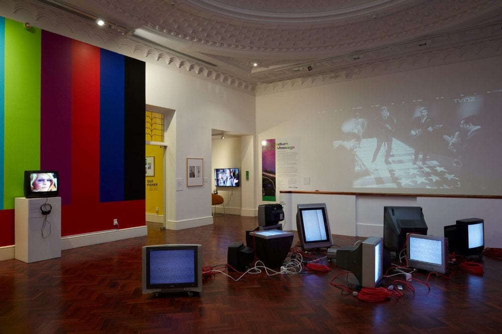 A gallery space with a colorful TV test pattern painted onto the left-hand wall, and a film projected on the right-hand wall. Several CRT television sets are scattered across the floor of the gallery.
