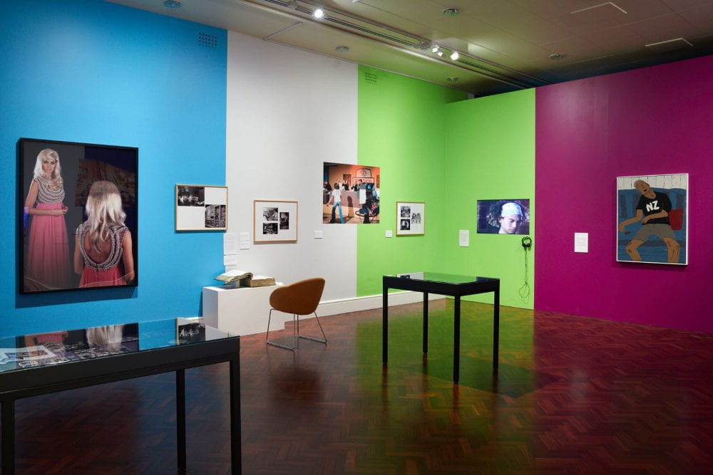 A gallery space with blue, white, green and pink walls. Various artworks are displayed throughout the space.